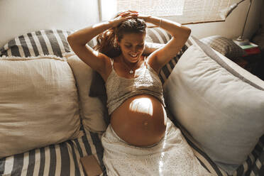 Glückliche schwangere Frau mit erhobenen Armen auf dem Sofa zu Hause sitzend - PCLF00728