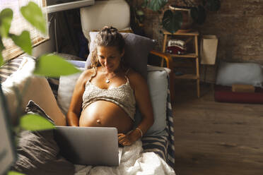Smiling pregnant woman using laptop on sofa at home - PCLF00730