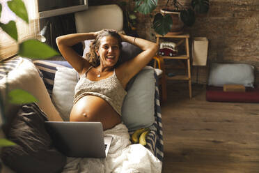Lächelnde schwangere Frau mit Laptop auf dem Sofa zu Hause - PCLF00731