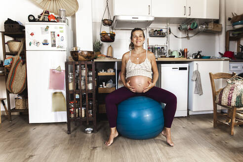 Lächelnde schwangere Frau, die in der Küche auf einem Fitnessball trainiert - PCLF00745