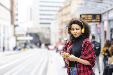 Smiling woman wearing headphones and holding smart phone in city - WPEF07683