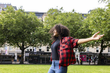 Smiling woman standing with arms outstretched in park - WPEF07696