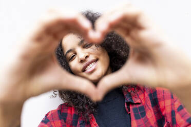 Smiling woman gesturing heart shape in front of wall - WPEF07711