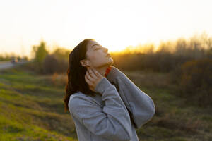 Porträt einer nachdenklichen Frau mit geschlossenen Augen auf einer Wiese bei Sonnenuntergang - TETF02290