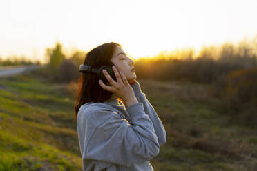 Seitenansicht einer Frau, die mit geschlossenen Augen auf einer Wiese bei Sonnenuntergang Musik hört - TETF02295