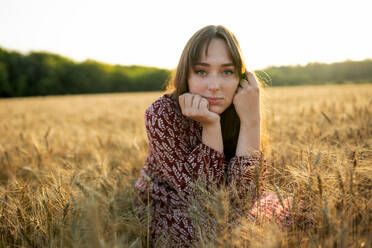 Portrait of young woman looking at camera while crouching in field at sunset - TETF02307