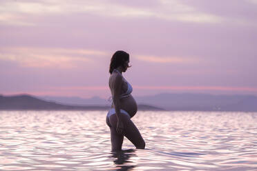 Side view of pregnant female in bikini with closed eyes while standing in water at beach against sunset sky in summer day - ADSF47889