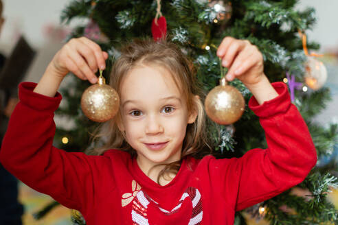 Lächelndes kleines Mädchen im roten Pullover, das in die Kamera schaut, während es Kugeln am Weihnachtsbaum zu Hause zeigt - ADSF47891