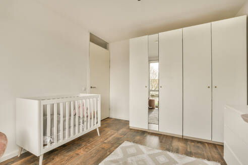 Kinderbett und Kleiderschrank aus Holz mit hellen Wänden und Parkettboden neben der Tür in einem stilvollen Babyzimmer in einem modernen Haus - ADSF47973