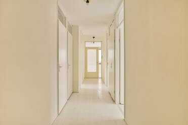 Interior of long narrow corridor leading to several rooms with white doors in modern house with beige walls and laminated floor - ADSF47983