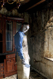 Anonymer furchterregender Mann im Metzgerkostüm mit Schürze und Messer schaut in die Kamera, während er in einem alten Wohnzimmer in der Nähe des Fensters steht - ADSF47995