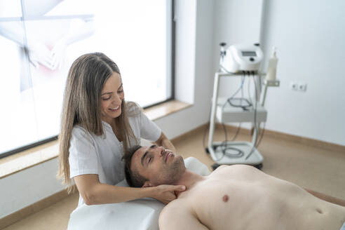 Young positive therapist in white uniform smiling and massaging the neck of a calm patient lying on a table shirtless during a physiotherapy session at a clinic - ADSF47996