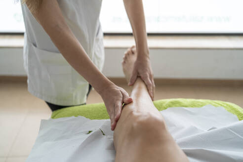 Unrecognizable therapist in uniform applying oil on leg of patient while preparing for massage session during rehabilitation process in modern clinic - ADSF48000