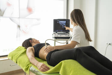 Professionelle weibliche Therapeutin untersucht den Bauch einer Patientin mit einem Ultraschallgerät, während sie ein Beratungsgespräch führt und auf einen Bildschirm in einer modernen Klinik zeigt - ADSF48004