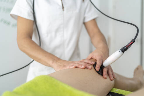 Unrecognizable professional doctor in uniform using shock wave therapy tool on knee of client during appointment in modern clinic - ADSF48006