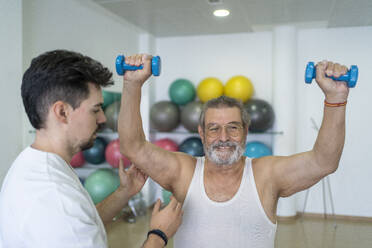 Seriöser männlicher Therapeut in Uniform hilft älteren Patienten bei Übungen mit Hanteln während des Rehabilitationsprozesses in einem modernen Klinikzentrum - ADSF48009