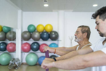 Serious male therapist in uniform helping senior patient to do exercise with elastic resistance band during rehabilitation treatment procedure in modern hospital - ADSF48010