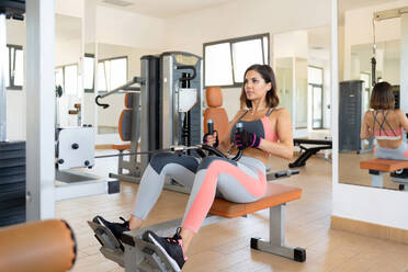 Full body of strong adult woman doing seated cable row on equipment in light gym - ADSF48017