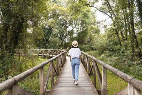 Woman wearing hat walking on footbridge in forest - MMPF00944