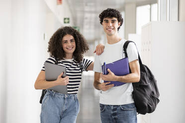 Happy students holding file folder and laptop in corridor - LMCF00608