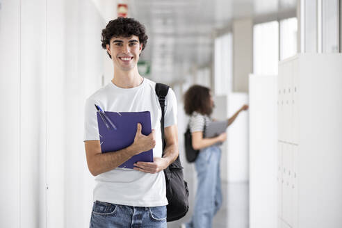 Happy student holding file folder and standing in corridor - LMCF00614