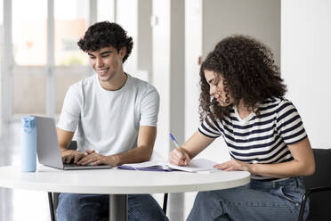 Smiling students writing on book and using laptop at table in university - LMCF00619