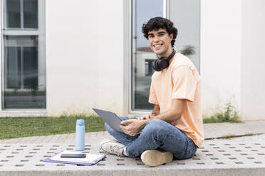 Lächelnder Student mit Laptop auf einer Betonbank auf dem Campus - LMCF00646