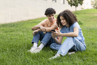 Smiling couple using smart phone on grass in park - LMCF00657