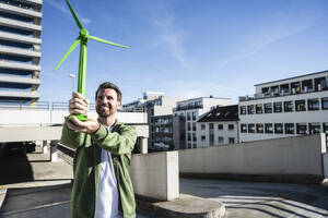 Lächelnder Ingenieur, der ein grünes Windradmodell auf einer Terrasse an einem sonnigen Tag hält - UUF30530