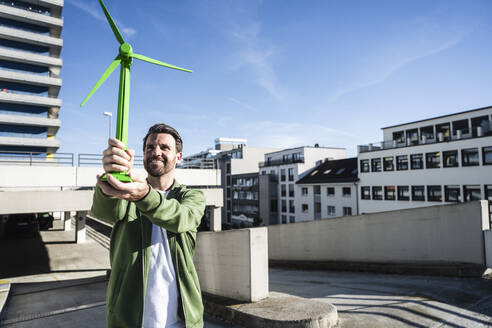 Smiling engineer holding green wind turbine model at terrace on sunny day - UUF30530