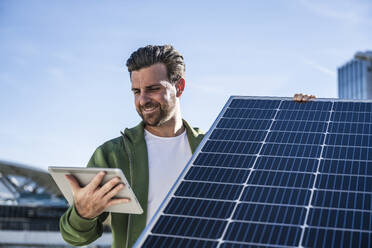 Lächelnder Ingenieur, der ein digitales Tablet und ein Solarpanel an einem sonnigen Tag hält - UUF30533