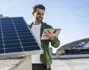 Lächelnder Ingenieur mit digitalem Tablet und blauem Solarmodul an einem sonnigen Tag - UUF30534