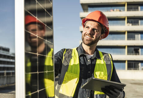 Engineer wearing protective workwear holding digital tablet examining solar panel on sunny day - UUF30561