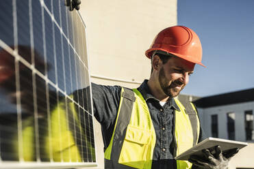 Lächelnder Ingenieur in Arbeitsschutzkleidung, der ein digitales Tablet mit einem Solarpanel benutzt - UUF30564