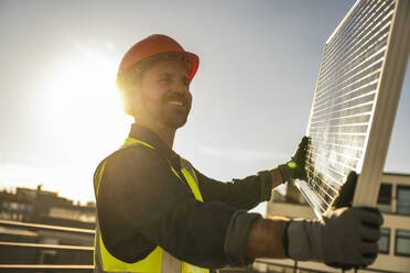 Von hinten beleuchteter lächelnder Ingenieur in Arbeitsschutzkleidung, der an einem sonnigen Tag ein Solarpanel hält - UUF30568