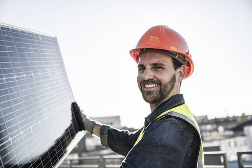 Lächelnder Ingenieur mit Schutzhelm, der ein Solarpanel hält - UUF30569