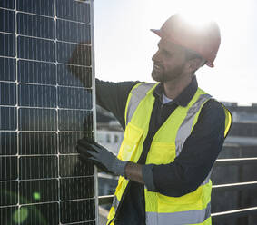 Von hinten beleuchteter Ingenieur, der an einem sonnigen Tag ein Solarmodul auf der Terrasse untersucht - UUF30572