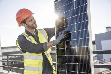 Lächelnder Ingenieur, der ein hintergrundbeleuchtetes Solarpanel auf einer Terrasse an einem sonnigen Tag untersucht - UUF30573