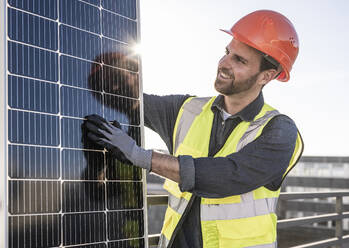 Lächelnder Ingenieur, der ein hintergrundbeleuchtetes Solarmodul auf einer Terrasse untersucht - UUF30574