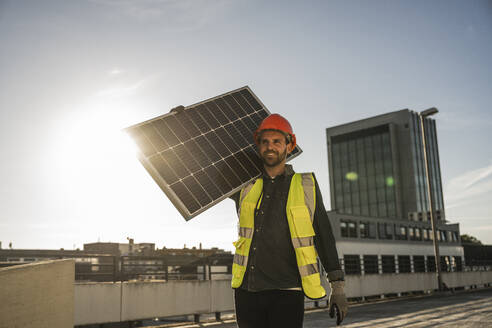 Ingenieur mit Schutzhelm trägt ein beleuchtetes Solarpanel auf der Schulter auf einer Terrasse - UUF30580