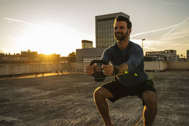 Lächelnder aktiver Mann, der auf der Terrasse mit einer Kettlebell trainiert - UUF30591