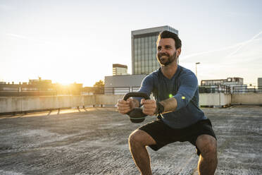 Lächelnder aktiver Mann in der Hocke mit Kettlebell auf der Terrasse - UUF30592