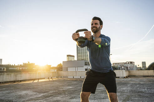 Smiling active man exercising with kettlebell at sunset - UUF30593