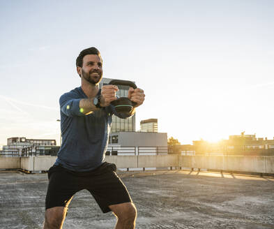 Lächelnder aktiver Mann, der auf der Terrasse mit einer Kettlebell trainiert - UUF30594