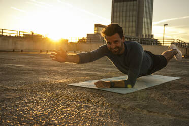 Smiling man practicing plank position on mat at sunset - UUF30599