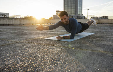Smiling active man practicing plank position on mat at sunset - UUF30600