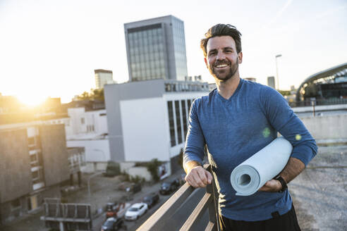 Happy man holding rolled up exercise mat leaning on railing at terrace - UUF30602