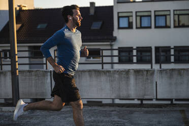 Aktiver Mann joggt in der Nähe des Geländers auf der Terrasse bei Sonnenuntergang - UUF30607