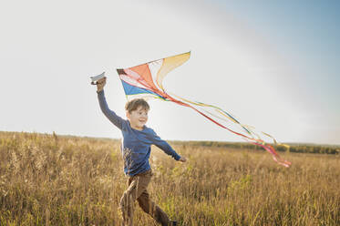 Lächelnder Junge lässt Drachen steigen und läuft im Feld unter dem Himmel - ANAF02149