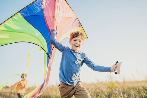 Smiling boy holding kite with brother running in field under sky - ANAF02168